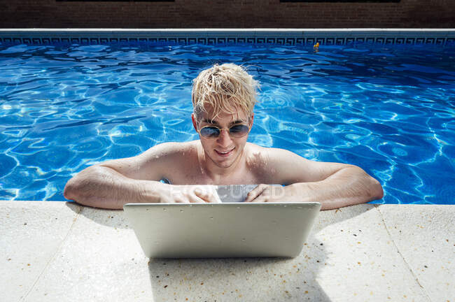Un hombre guapo sin camisa usando gafas de sol usando un portátil junto a la piscina - foto de stock