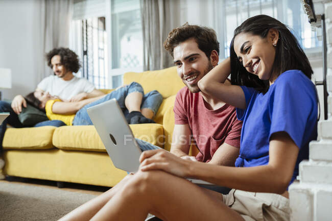 Lächelndes junges Paar teilt Laptop, während Freunde es sich zu Hause auf dem Sofa gemütlich machen — Stockfoto