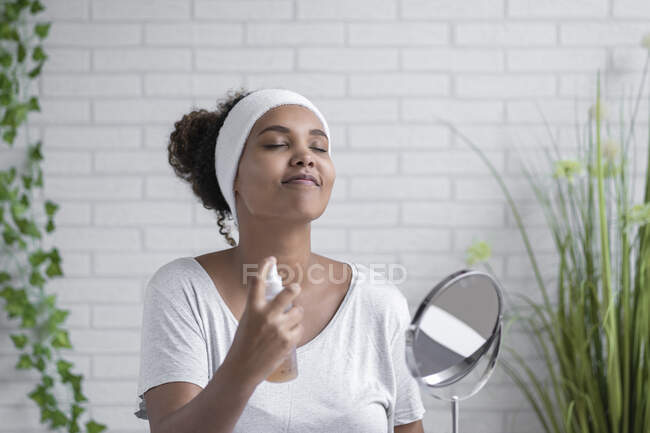 Giovane donna con gli occhi chiusi spruzzando nebbia facciale a casa — Foto stock