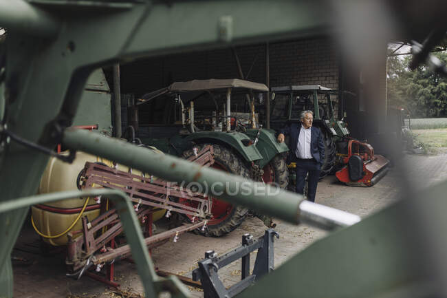 Empresário sénior numa quinta com tractor no celeiro — Fotografia de Stock