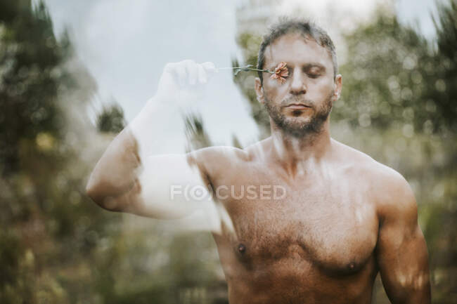 Shirtless bonito homem com olhos fechados segurando flor no olho na floresta visto através de vidro — Fotografia de Stock