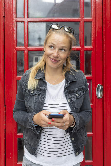 Smiling woman holding smart phone looking away while standing against telephone booth — Stock Photo