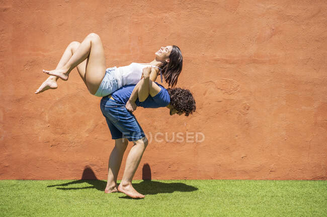 Hombre joven llevando a la mujer en la espalda por la pared marrón durante el día soleado - foto de stock