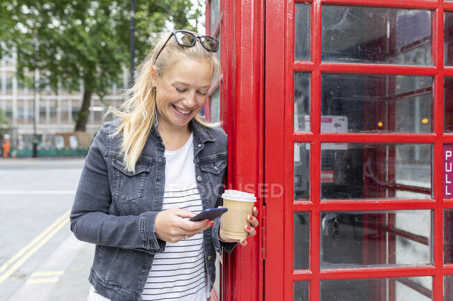 Smiling woman holding coffee cup using smart phone while standing by telephone booth — Stock Photo