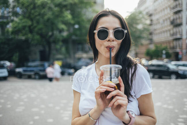 Young woman wearing sunglasses drinking soft drink while standing in city — Fotografia de Stock