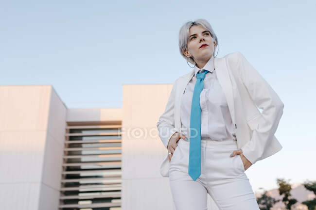Молода жінка в білому костюмі позує, стоячи на тлі чистого неба в місті — стокове фото