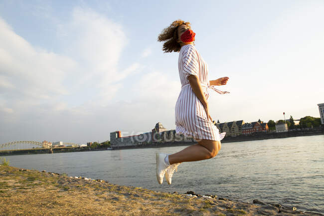 Mitte erwachsene Frau mit Maske springt am Flussufer gegen den Himmel in der Stadt — Stockfoto
