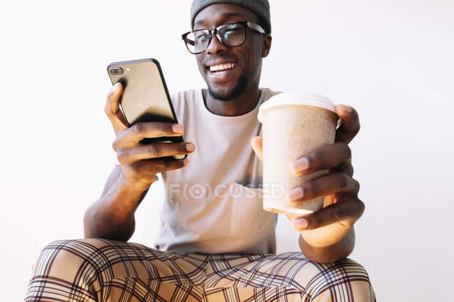 Sonriente joven usando el teléfono inteligente mientras está sentado con la taza de café desechable contra el fondo blanco - foto de stock