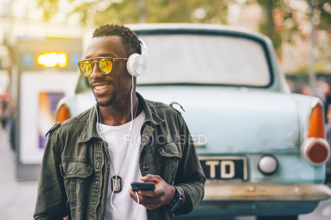 Smiling young man wearing sunglasses listening music through headphones against vintage car in city — Stock Photo