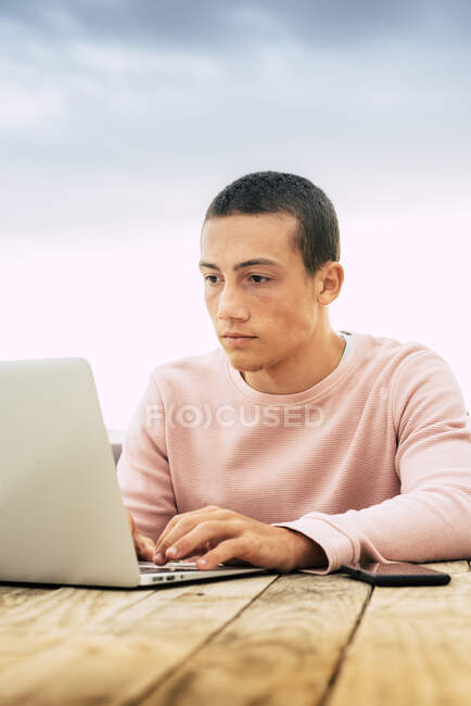 Menino adolescente com cabelo curto usando laptop na mesa contra o céu — Fotografia de Stock