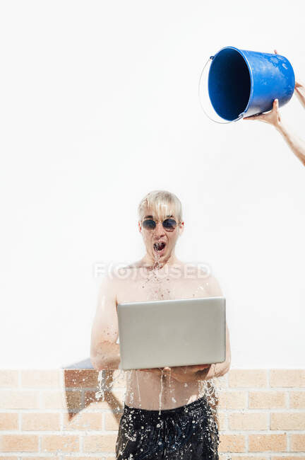 Mediados de la mujer adulta vertiendo agua con cubo en el hombre joven sin camisa utilizando el ordenador portátil contra la pared - foto de stock