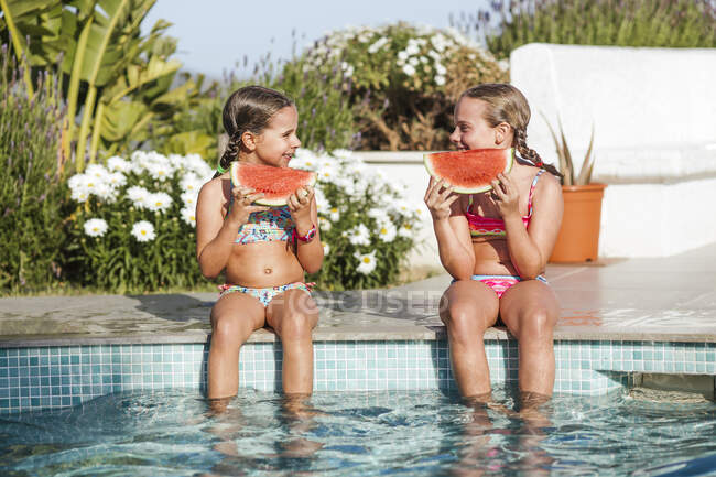 Chicas gemelas sonrientes sentadas con rebanada de sandía en la piscina en un día soleado - foto de stock