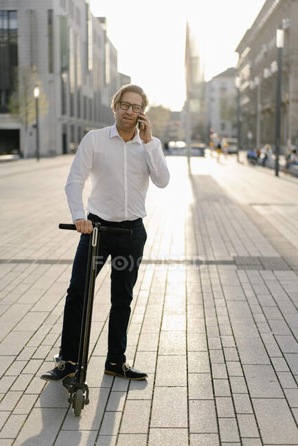 Businessman with kick scooter on the phone in the city — Stock Photo
