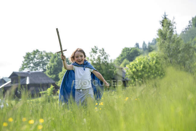 Играющий мальчик в плаще с игрушечным мечом, бегущий по травянистой земле под ясным небом — стоковое фото