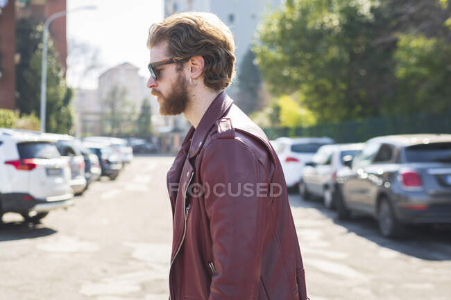 Молодой человек в солнцезащитных очках и кожаной куртке идет по дороге по городу — стоковое фото
