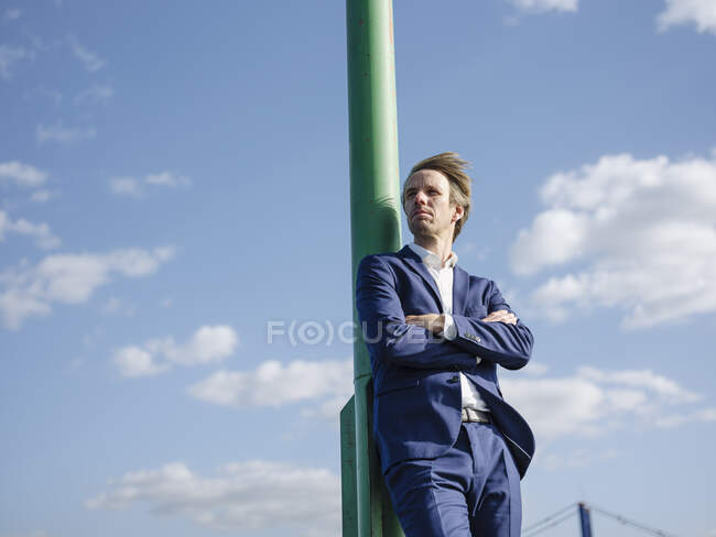 Pensativo hombre de negocios con los brazos cruzados de pie por poste contra el cielo durante el día soleado - foto de stock