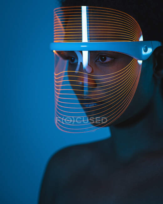 Primer plano de la mujer que usa máscara de terapia de luz azul contra la pared en casa - foto de stock