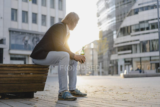 Бізнесмен сидить на лавці в місті під час заходу сонця з пляшкою пива. — стокове фото