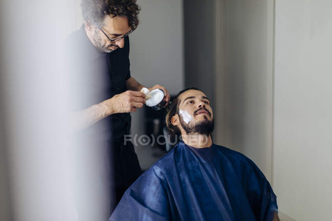 Barber applying shaving cream on client in salon — Stock Photo