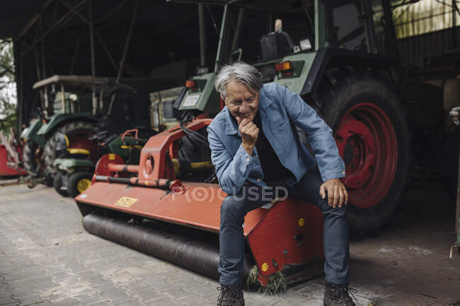 Улыбающийся пожилой человек на ферме сидит на тракторе в сарае — стоковое фото