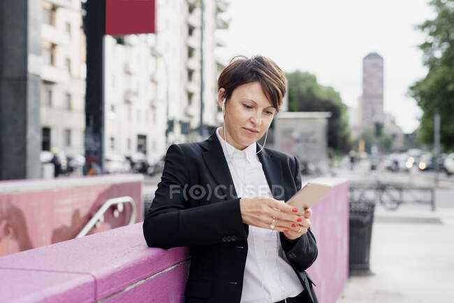 Confident businesswoman using smart phone while standing by retaining wall in city — Foto stock