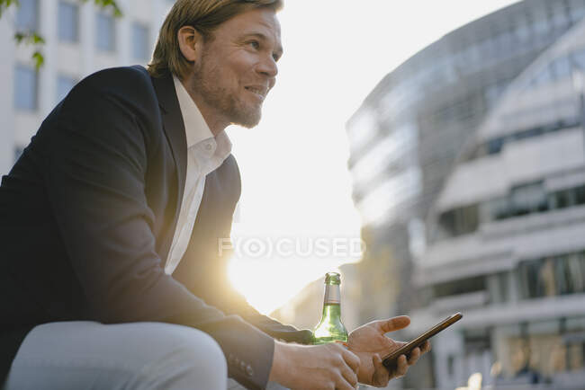 Uomo d'affari seduto su una panchina in città al tramonto con una bottiglia di birra e smartphone — Foto stock
