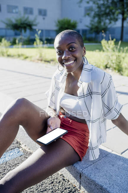 Посмішка молодої жінки з голеною головою за допомогою мобільного телефону, сидячи на стежці в місті. — стокове фото