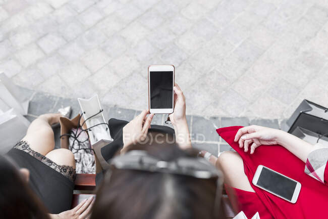 Mujer joven usando el teléfono inteligente mientras está sentada con amigas en el banco de la ciudad - foto de stock