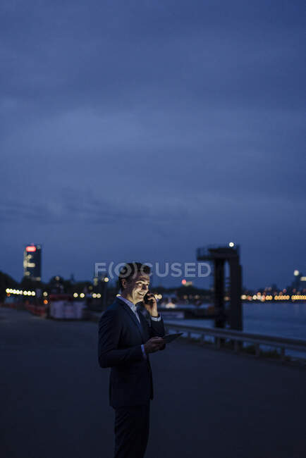 Reifer Geschäftsmann mit Tablet und Smartphone in der Abenddämmerung am Flussufer — Stockfoto