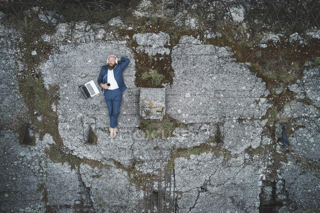 Vue aérienne d'un homme d'affaires portant un costume écoutant de la musique alors qu'il était allongé sur la terre ferme en forêt — Photo de stock
