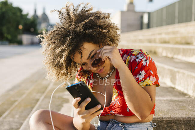 Trendy woman using smart phone while sitting on steps in city — Stock Photo