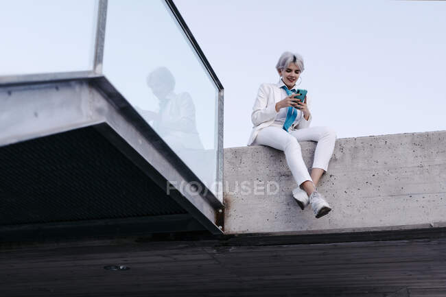 Молода жінка в білому костюмі, використовуючи смартфон, сидячи на тримаючій стіні в місті — стокове фото