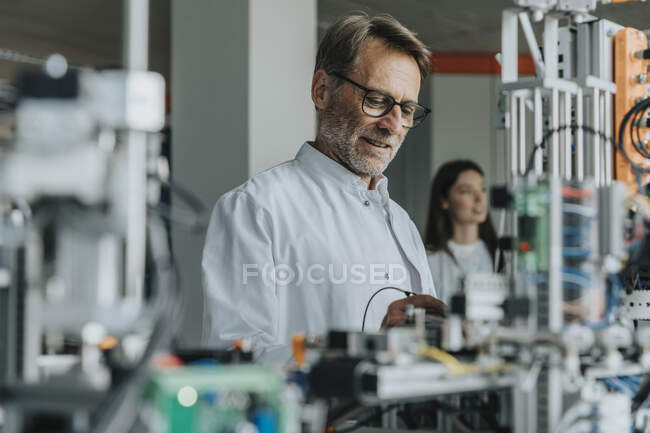 Uomo maturo esaminando macchinari con collega di sesso femminile in background in laboratorio — Foto stock