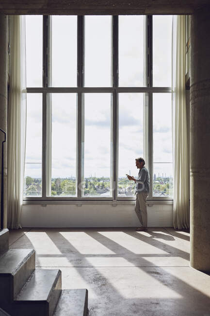 Senior mit Smartphone am Fenster einer Dachgeschosswohnung — Stockfoto