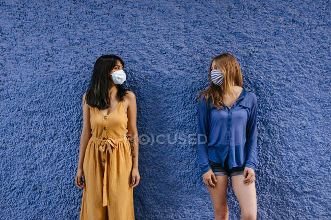 Le amiche che indossano maschere che si guardano mentre mantengono la distanza contro il muro blu in città — Foto stock