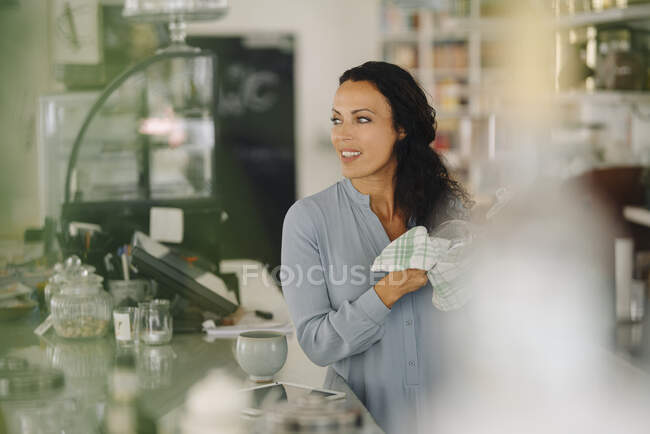 Femme propriétaire tenant serviette regardant loin tout en se tenant au comptoir dans un café — Photo de stock