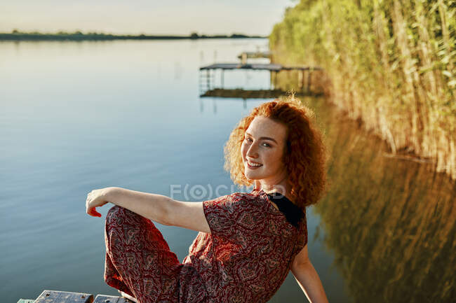 Retrato de sorridente ruiva jovem sentada no molhe em um lago ao pôr-do-sol — Fotografia de Stock