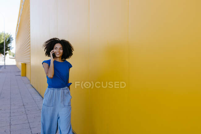 Mujer joven hablando por teléfono, caminando por la pared amarilla - foto de stock