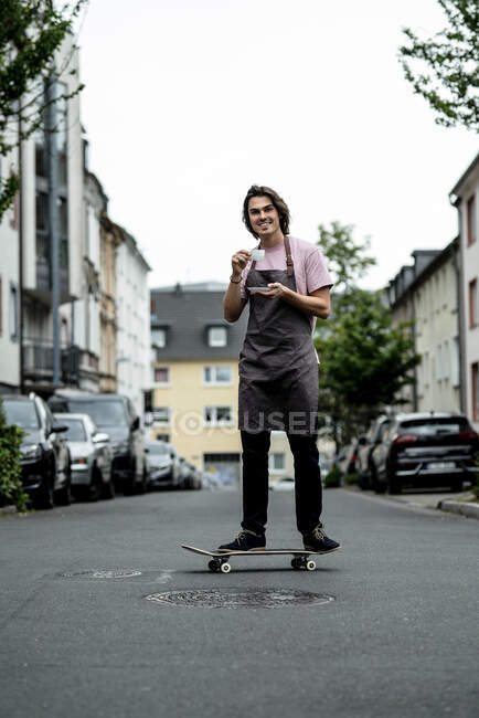 Smiling male owner holding coffee cup while skateboarding on road in city — Stock Photo