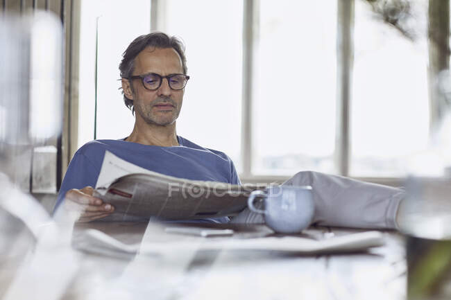 Senior man sitting at the table in a loft flat reading newspaper — Stock Photo