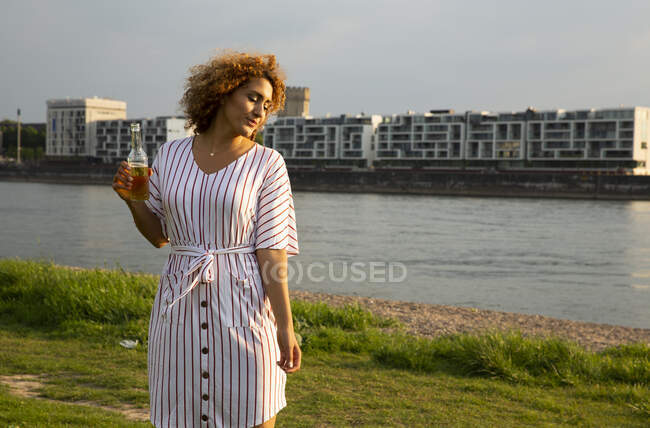 Mid adult woman holding beer bottle standing on grassy land against river in city at sunset — Stock Photo