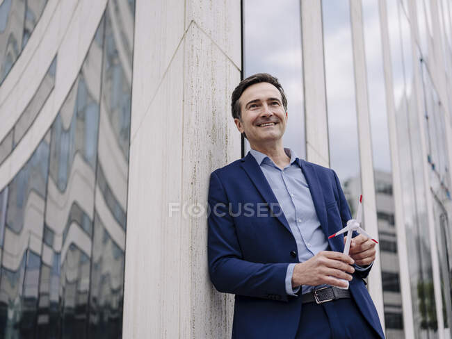 Hombre de negocios maduro sonriente con modelo de turbina eólica apoyado en un edificio de la ciudad - foto de stock