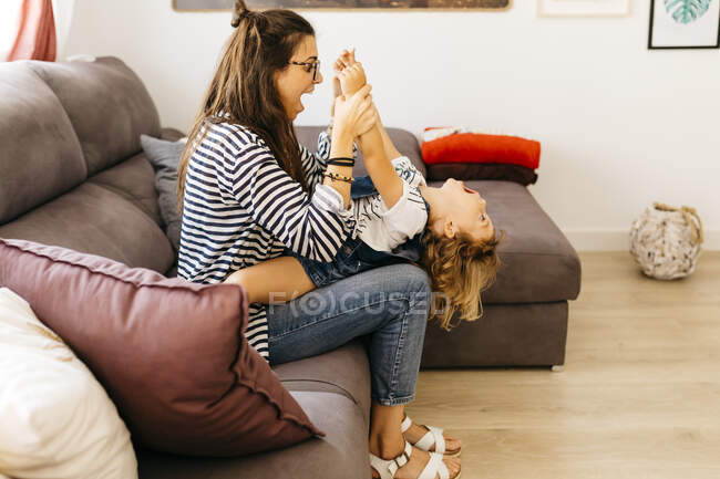 Madre con la boca abierta sosteniendo las manos de la hija juguetona mientras está sentada con ella en el sofá en casa - foto de stock