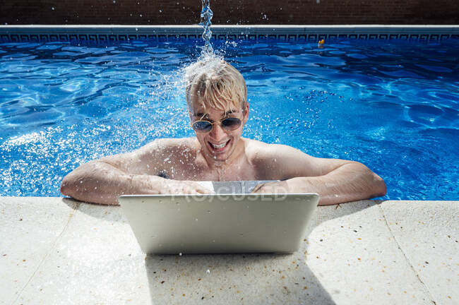 Wasser spritzt auf hemdslosen jungen Mann mit Laptop am Pool — Stockfoto