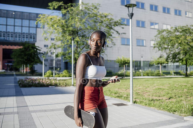 Young woman holding mobile phone and skateboard looking away while standing on footpath in city — Stock Photo