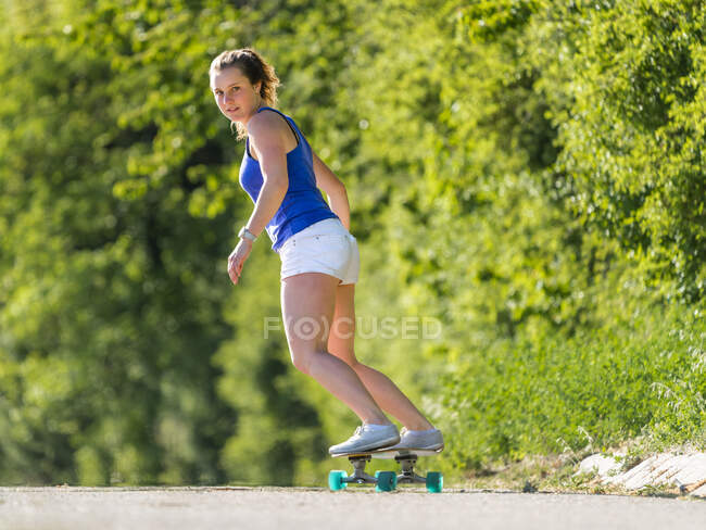 Giovane donna skateboard su strada dalle piante durante la giornata di sole — Foto stock