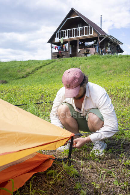 Homem adulto médio vestindo tampa instalar barraca em terras gramadas no acampamento contra o céu — Fotografia de Stock