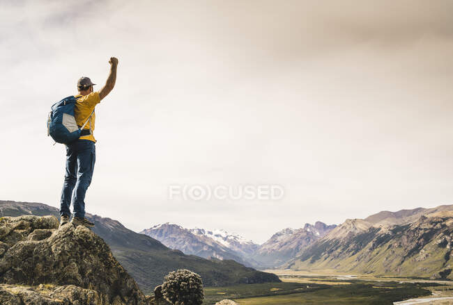 Hombre con brazo levantado mirando montañas mientras está de pie sobre roca, Patagonia, Argentina - foto de stock