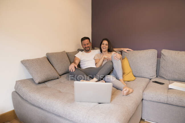 Feliz pareja viendo la película sobre el ordenador portátil mientras se relaja en el sofá en casa - foto de stock