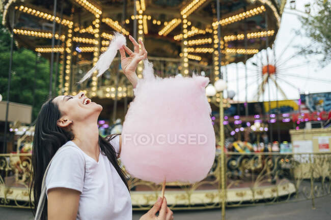 Happy beautiful woman eating cotton candy while standing against carousel in amusement park — Fotografia de Stock
